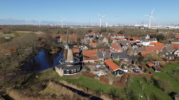 Uitzicht op een dorp met huizen, bomen en een waterloop, omringd door windturbines.